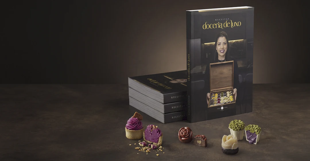 O livro Receitas Doceria de Luxo em pé sobre a bancada ao lado de exemplares empilhados e uma fileira de doces finos, com Natália Canezin na capa segurando uma caixa de bombons.